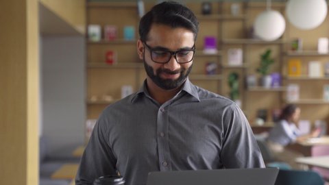 Happy indian businessman holding coffee and laptop walking in office. Smiling male employee, business man executive going along modern office coworking space using computer.