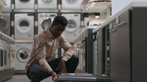 adult afro-american is choosing dishwasher in home appliance store, opening door and examining inside
