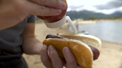squeezing ketchup on a hot dog or bratwurst on the beach of a lake in the mountains.