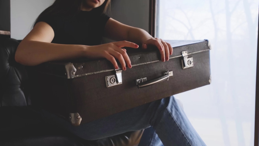 A young woman sits on the couch and holds an old suitcase on her knees. Fingers rattling on the suitcase. Window, bright light. Concept of waiting for traffic, train station.