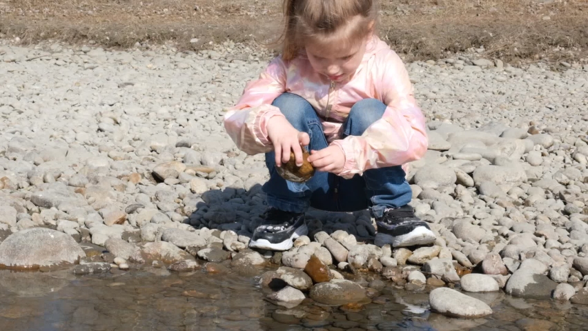 Close-up of caucasian child girl standing on stones rock in natural stream in springtime or autumn  and playing with stones. investigate  details of nature . outdoor kids activity and learning concept