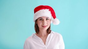 Beautiful smiling caucasian woman wearing white shirt and red santa hat shows thumbs up sign. Blue background. Studio shot. 4K resolution video. Positive emotion theme.