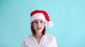 Beautiful smiling caucasian woman wearing white shirt and red santa hat shows good idea hand sign. Blue background. Studio shot. 4K resolution video. Positive emotion theme.