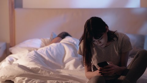 Internet anxiety. Night insomnia. Social media. Disturbed woman using mobile phone sitting on bed with sleeping husband late in home bedroom.