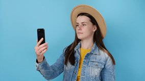 Cheerful cute young woman talking on mobile phone making video call greeting with hand point finger showing thumb up, wears denim jacket and hat, isolated on blue studio wall. People lifestyle concept