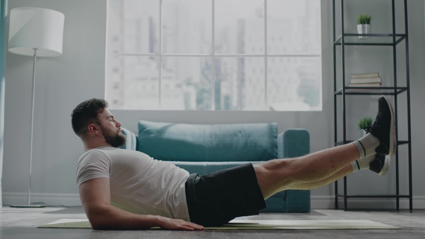 Muscular athlete with beard practices flutter kicks exercise lying on yellow fitness mat at home in early morning slow motion