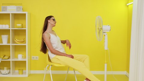 Young stylish woman, model with long straight hair, is posing on chair in front of electric fan that is blowing her long hair, she is trying to cool down and relax in hot office, Slow motion.