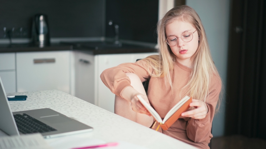 Beautiful 10 years old blond girl in glasses sitting at the desk and reading a book while doing her homework.