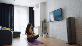 Young woman with ponytail wearing sports clothes sitting in lotus posture in front of TV screen and repeating after instructor streaming from seaside. Practicing yoga at home. Concept of fitness
