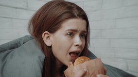 Comfort Eating. Depressed Young Woman Eats Burger Feeling Sad Sitting In Bed Wrapped In Blanket, Suffering From Depression At Home. Female Biting Cheeseburger Having Bulimia Disorder, Zoom In