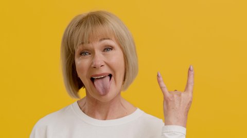 Mature rock lover. Playful positive middle aged lady showing punk gesture and sticking tongue out, feeling excited, orange studio background, slow motion