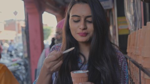 front view shot of a young beautiful Indian woman enjoying the street food while drinking a cold beverage drink called Lassi in an earthen clay pot at an urban market place