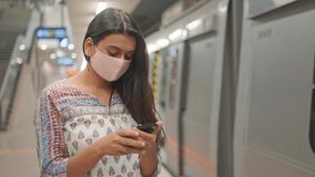 A mid-shot of a young beautiful Indian Asian female wearing a protective face mask standing and using a mobile phone on a platform next to a city metro train or subway amid COVID 19 epidemic 