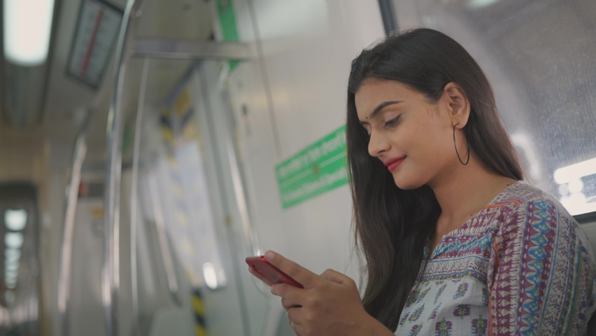 Close shot of a young attractive Indian female in traditional salwar kurta sitting on a moving city metro train and typing a text message on a mobile phone  Royalty-Free Stock Footage #1071069292