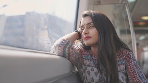 A close shot of a beautiful Indian young woman solo traveler in traditional salwar kurta sitting in a moving metro train leaning on a window and looking outside 