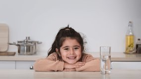 Portrait Of Cute Arabic Little Girl Posing With Glass Of Water And Smiling To Camera Sitting In Modern Kitchen At Home. Healthy Liquid. Kid's Headshot For Mineral Aqua Advertisement. Zoom In Shot
