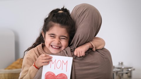 Mother's Day Holiday. Muslim Daughter Hugging Mommy In Hijab Holding Darwn Greeting Card With Heart Smiling To Camera Sitting In Kitchen At Home. Cute Kid Girl Embracing Mom Expressing Love Indoors