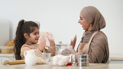 Happy Muslim Mother In Hijab And Little Daughter Having Fun With Flour Giving High-Five Baking Pastry In Modern Kitchen At Home. Young Arabic Mom And Her Kid Girl Cooking Cake Together