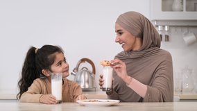 Happy Muslim Mother Feeding Daughter Having Lunch Together Drinking Milk And Eating Cookies Sitting In Modern Kitchen On Weekend. Islamic Family Nutrition Concept. Slow Motion