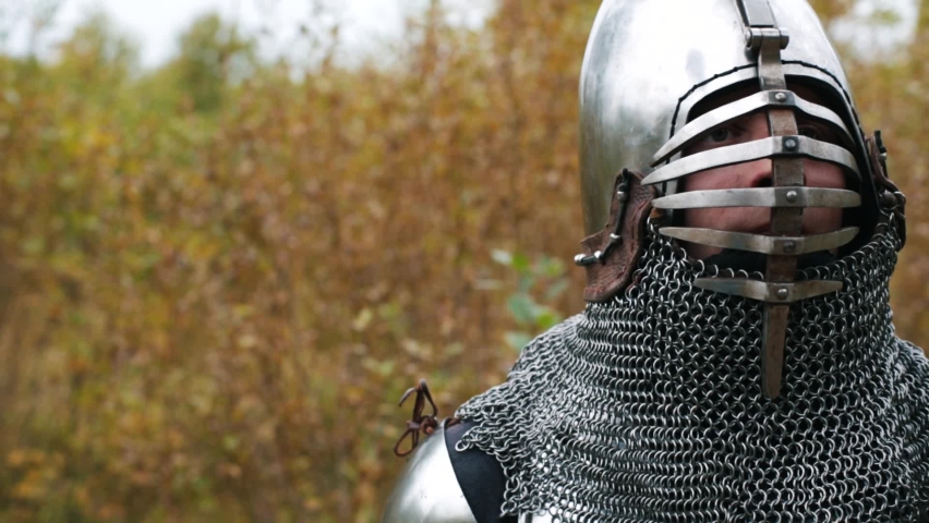 Close-up of a medieval knight in armor, who removes his helmet from his head, stares into the distance, grins, stretches his arms and walks out of the frame against the background of autumn trees.