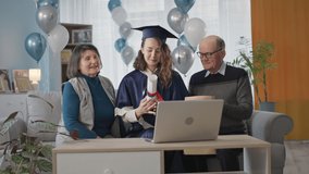young woman in mantle holding diploma with her parents celebrating end of school year online during an online ceremony using video link on laptop