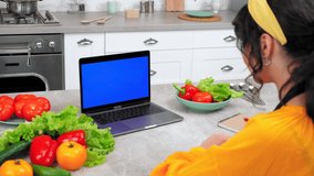 Blue screen mock up chroma key monitor laptop concept: Woman housewife in kitchen watch online cooking course listen teacher chef writes culinary recipe in notebook, study remote video call computer