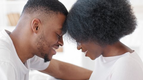Loving young african couple bonding laughing smiling touching foreheads together looking at camera, romantic millennial afro man and happy black woman getting closer enjoying affection moment at home