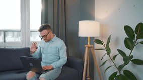 Casual dressed man with glasses sitting on the couch and using laptop to make a video call from home in a modern apartment