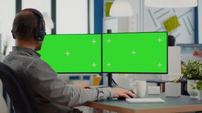 Employee with headphones using dual monitor setup with green screen, chroma key mock up isolated display sitting in video production studio. Man editor processing film montage on pc in creative agency