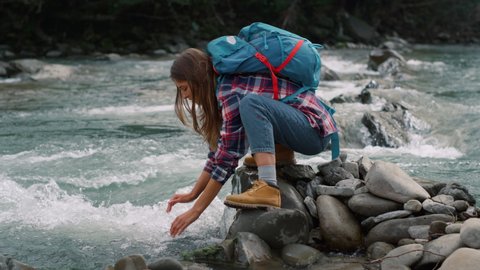 Female tourist touching water in river. Tired woman with backpack drinking fresh water from mountain stream. Attractive girl washing face with water from river during hike