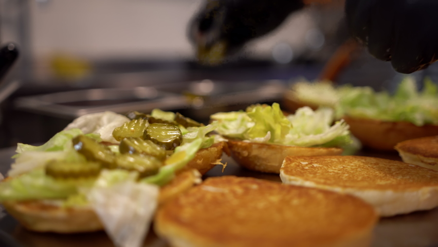 Preparing burgers in a fast food restaurant. Close up view of a fast food worker adding vegetables to the burger Royalty-Free Stock Footage #1071087988