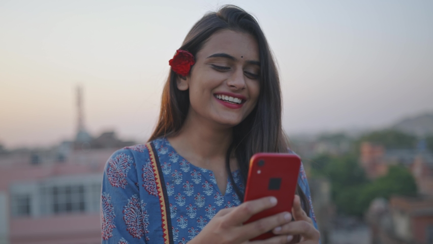 A young Indian Asian cheerful attractive woman is holding a mobile phone in hands and watching a comedy video or movie on an online stream or OTT platform smiling and standing outdoors in the evening Royalty-Free Stock Footage #1071088228
