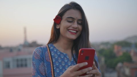 A young Indian Asian cheerful attractive woman is holding a mobile phone in hands and watching a comedy video or movie on an online stream or OTT platform smiling and standing outdoors in the evening