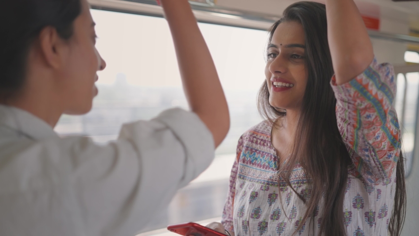 A close shot of a happy and beautiful young Indian woman in traditional dress holding a mobile phone in her hand standing and having a conversation with a fellow passenger in a moving metro train Royalty-Free Stock Footage #1071088246