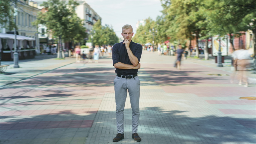 Time lapse of attractive blond guy standing outdoors in busy street with pensive expression and looking at camera while people are rushing around