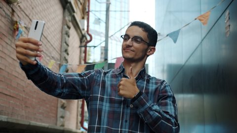 Slow motion of cheerful Middle Eastern man taking selfie with thumbs-up gesture using smartphone camera outdoors in city street. People and photography concept.