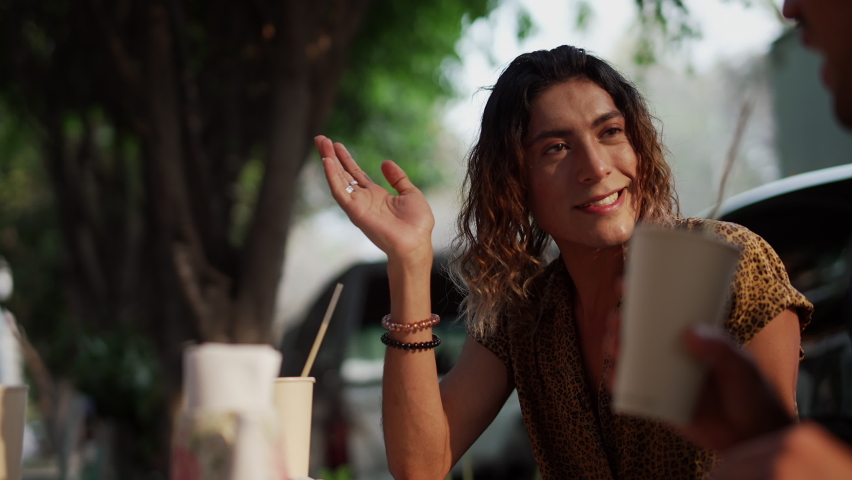 Young gay man with long hair in a printed t-shirt talking to his friends while sitting at the table of a cafe in Mexico City. Having a frappe. Looking entertained in the conversation. 