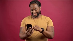 Young African-American man standing isolated on red wall holding mobile phone and scrolling news feed, watching posts in social media online, using dating app, enjoying new application