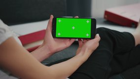 Young Woman sitting on sofa while looking at movies using phone in horizontal mode with mock up green screen chroma key display. Girl Using Mobile Phone, Watching Content, Videos, Blogs, Browsing Inte