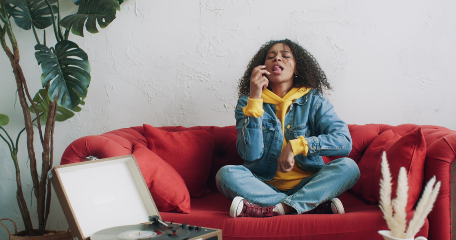 Beautiful african american woman in yellow hoodie and jeans jacket having fun listening music on vinyl turntable vintage record player dancing sitting on red sofa at home. Gen z cheerful urban female Royalty-Free Stock Footage #1071100384