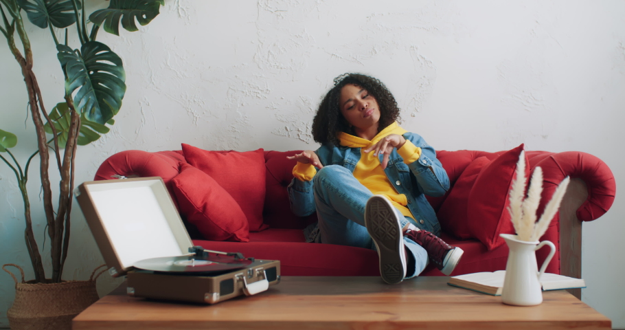 Happy stylish african american black female in yellow hoodie and jeans jacket listening music on vinyl turntable vintage record player dancing sitting on red sofa at home. Gen z cheerful urban woman Royalty-Free Stock Footage #1071100390