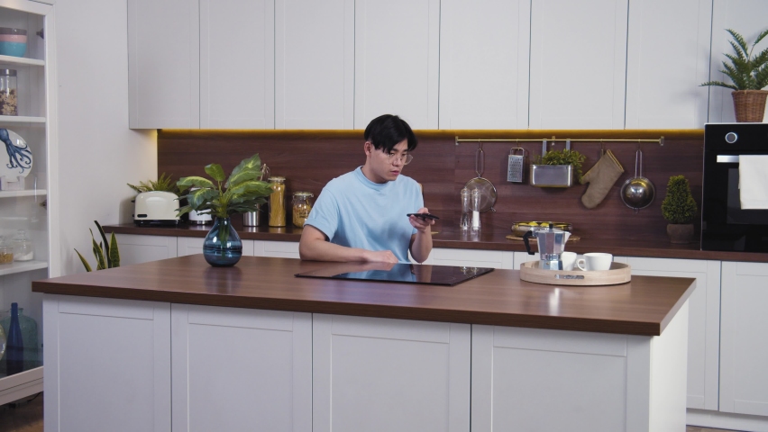 Smiling vietnamese man recording voice message at home kitchen. Happy young guy use phone for social network and voice control. General shot, slow motion 4k
