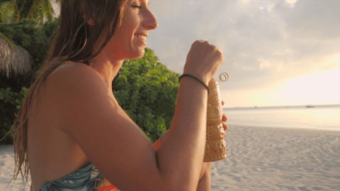 Slow motion: Young female enjoys sunset with a cocktail from tropical beach bar. Woman on vacation having a drink on the beach 