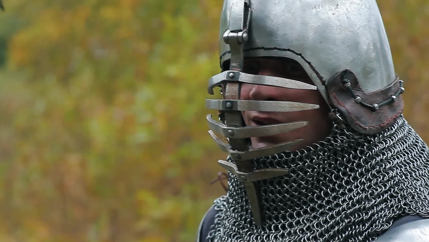A medieval knight in armor removes his helmet from his head, sweat dripping from his tired face. Close-up of the warrior against the background of yellowed autumn trees.