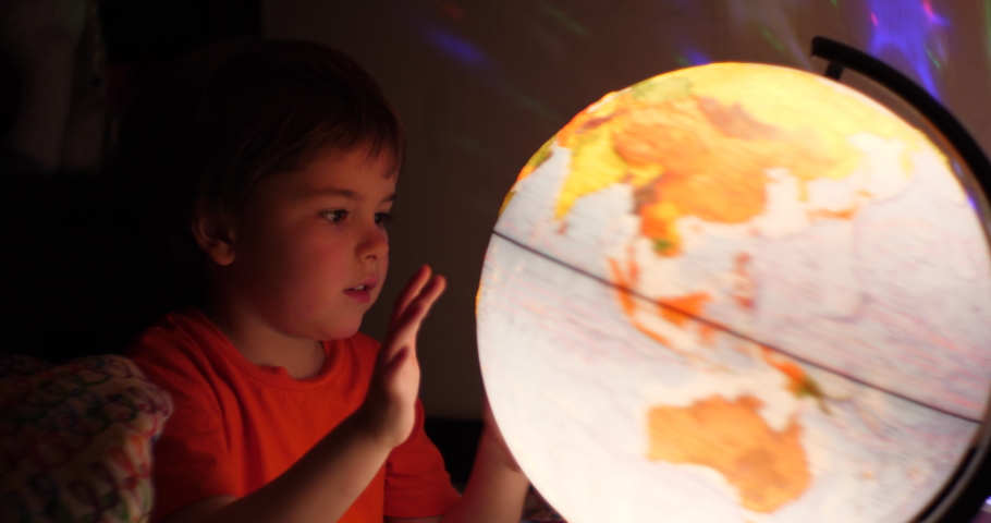 Boy Playing With Globe at Night. Child Sits on Bed in Evening Light Dreaming Vacation. Little Child Looking at Illuminated Globe, Exploring World, Learning. Dreaming About Future Save of Our Planet.