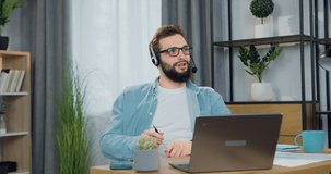 Good-looking cheerful relaxed 30-aged bearded guy in headset sitting in front of computer during video call with friend and enjoying conversation