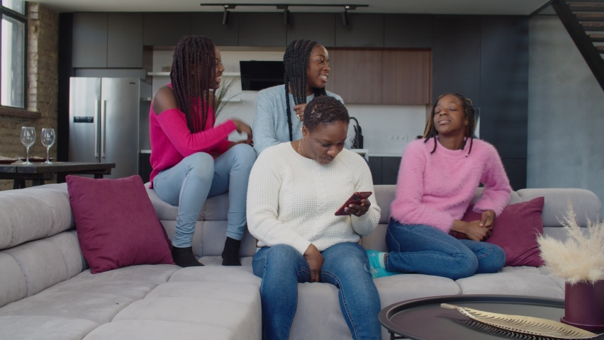 Strict attractive african mother scolding and calming down mischievous cute adolescent daughters fooling around on couch, making business call on mobile phone while family resting on couch indoors. Royalty-Free Stock Footage #1071137020