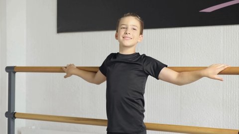 Portrait of a young handsome guy, in a black T-shirt and shorts, looks at the camera and smiles. Training at a ballet school. Hands on the barre.