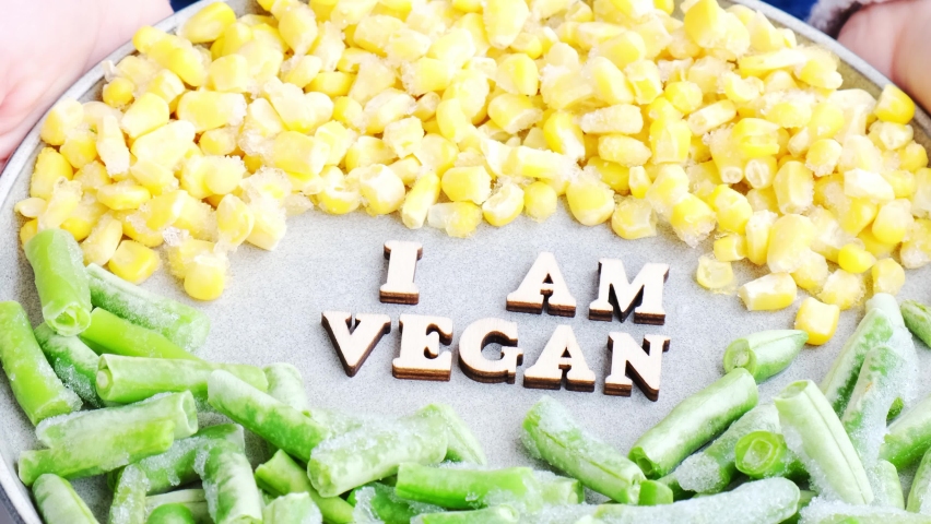 Female hands hold I AM VEGAN text in plate. Veganism, vegetarian healthy lifestyle green beans yellow corn. Healthy eating vegan. Wooden letters