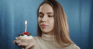 Portrait of young woman blowing out the candle on delicious sweet cake with berries, looking in camera with wide smile. Close-up of female student with green eyes. Happy beautiful model in studio.